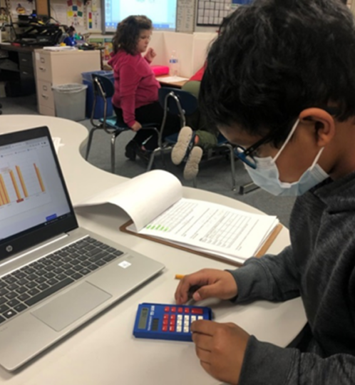 Masked student in a hoodie sits at a desk with a laptop, calculator, and notebook, studying as bar charts appear on the laptop screen in a classroom.