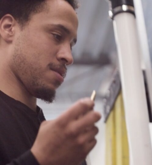 Person with short hair and light beard in profile, holding a small object between their fingers and looking at it, with a blurred outdoor urban background.