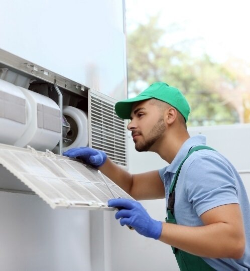 An outdoor HVAC technician wearing a green cap and blue gloves inspects a filter on an air conditioning unit.