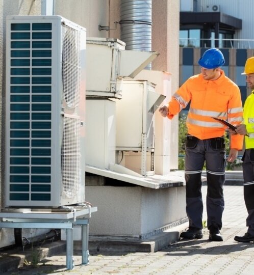 Two workers in high-visibility safety jackets and hard hats inspect a rooftop HVAC unit beside other industrial equipment on a paved area.