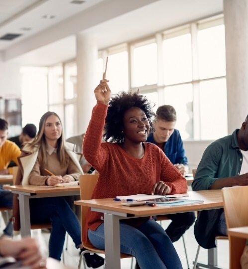 Person with curly hair wearing an orange sweater raises their hand with a pen in a bright classroom, as classmates sit at desks taking notes.