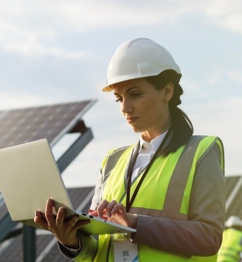 Person in a white hard hat and neon hi-vis vest uses a laptop outdoors near solar panels.