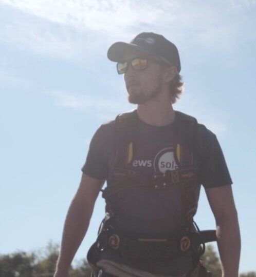Person wearing a cap, sunglasses, and climbing harness stands outdoors against a clear blue sky.
