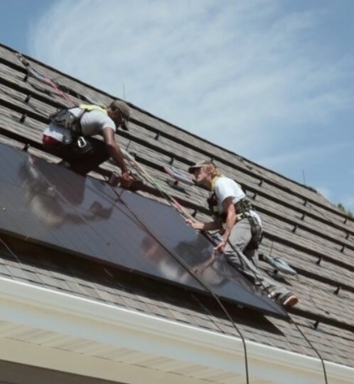 Two workers on a sloped roof installing a dark solar panel array, wearing safety harnesses, with a blue sky above.