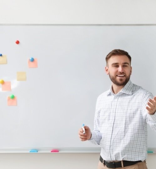 A bearded person in a light checkered shirt stands before a whiteboard with colorful sticky notes, holding a marker and gesturing as they speak.
