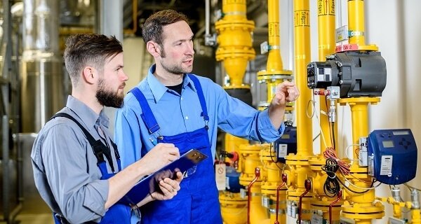 Two workers in blue overalls inspect yellow industrial pipes and gauges; one holds a tablet while the other points at a control valve.