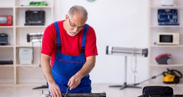 Person with gray hair in a red shirt and blue overalls, working on an electronic device at a workbench in a bright workshop.