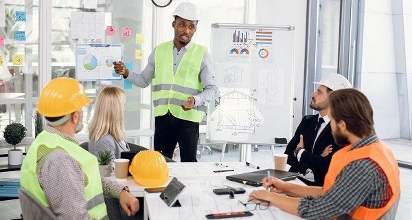 Person in a white hard hat and green safety vest presents project plans to a diverse group around a conference table, with charts on a whiteboard nearby.