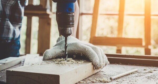 Person in a plaid shirt drills into a wooden block on a workbench; a glove rests on the block amid sawdust as sunlight streams through the window.