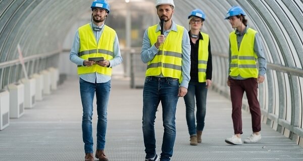 Four workers in high-visibility vests walk through a curved glass corridor; one speaks into a microphone while another holds a tablet.