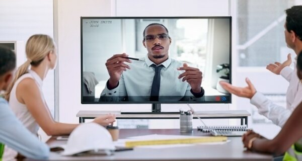 Diverse group seated around a conference table watches a presenter on a screen delivering a presentation; safety gear like hard hats rests on the table.