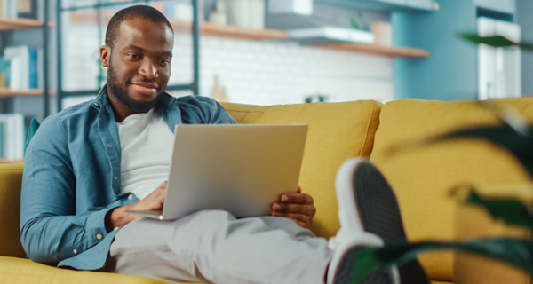 Bearded person lounging on a yellow sofa, using a laptop in a bright, modern space with teal cabinets and pendant lights.