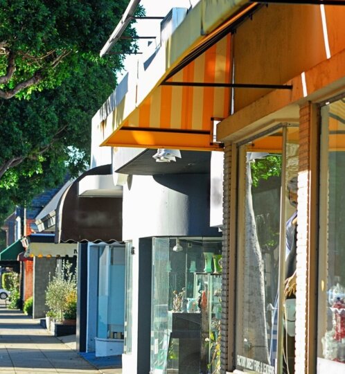 Sunny urban street with storefronts and orange striped awnings, glass display windows, and tall green trees along a shaded sidewalk.