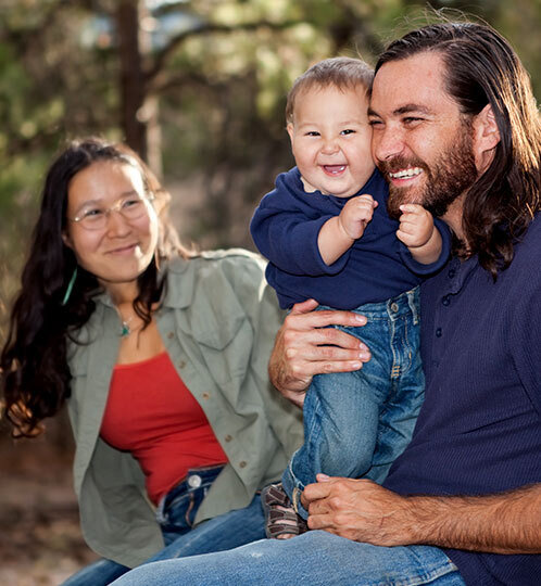 Three people outdoors in a park: a bearded adult holds a smiling baby in blue, while another adult with glasses sits nearby.
