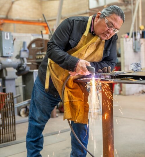 A worker in safety glasses and a brown leather apron grinds metal on a table, sparks flying in a busy workshop.