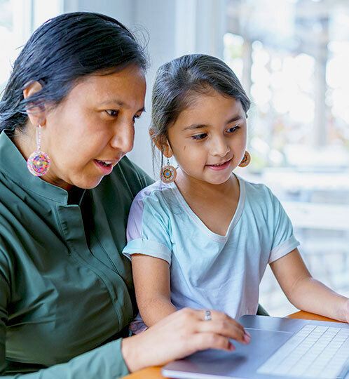 An adult and a child sit side by side at a table, looking at a laptop together in a bright indoor setting.