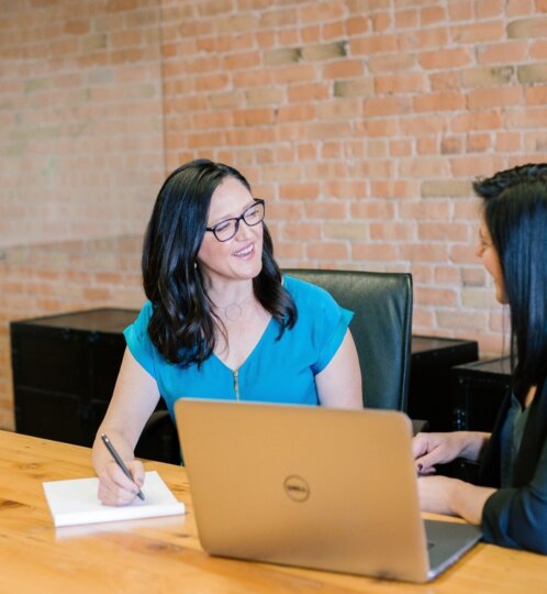 Two people sit at a wooden table in a brick-walled room; one in a blue blouse writes in notebook, the other in a dark blazer uses a gold Dell laptop.