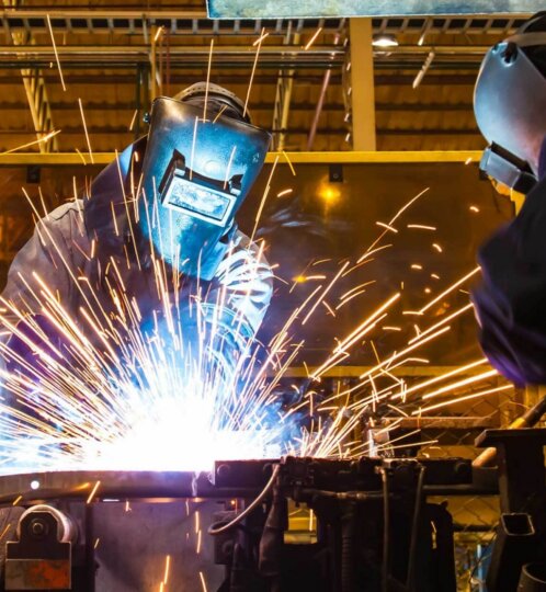 Two workers in protective welding gear and helmets weld metal on a bench as bright sparks fly in an industrial workshop.