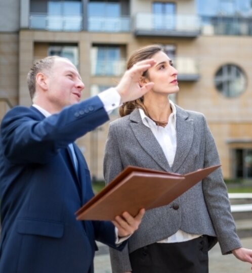 Two professionals outside a modern building; one in a blue suit points with a folder, other in a gray blazer shields eyes while looking ahead.