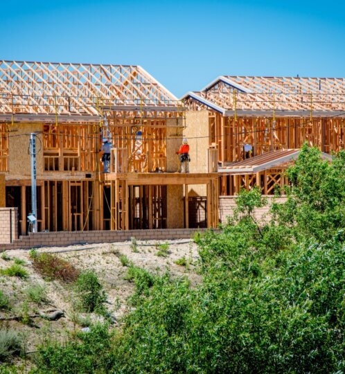 Two-story wooden-framed houses under construction with workers on scaffolding, a bright blue sky, and green shrubs in the foreground.