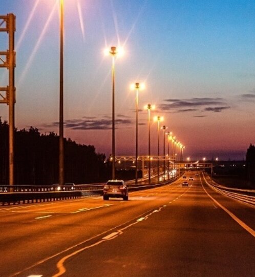 Highway at dusk with tall streetlights along a curved road, several cars, and tree-lined shoulders under a blue-to-orange sunset sky.