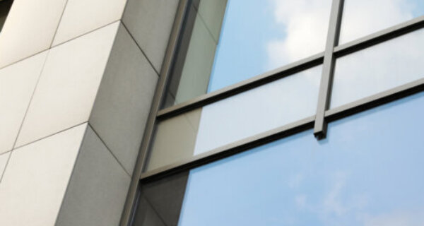 Close-up of a modern building facade with large reflective glass panels and light gray stone sections, reflecting a blue sky with clouds.