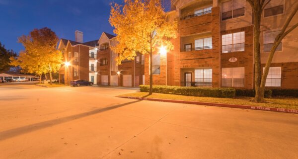 Evening view of a brick apartment complex with warm streetlights, yellow autumn trees, and a paved parking lot with a few parked cars.