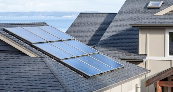 Sloped gray roof with two rows of blue solar panels, beige siding, and a wooden porch beam; distant ocean and shoreline visible.