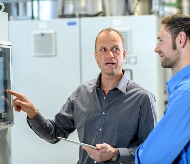 Two professionals in industrial setting discuss a touchscreen panel; one points at the screen while holding a tablet, with machinery in background.