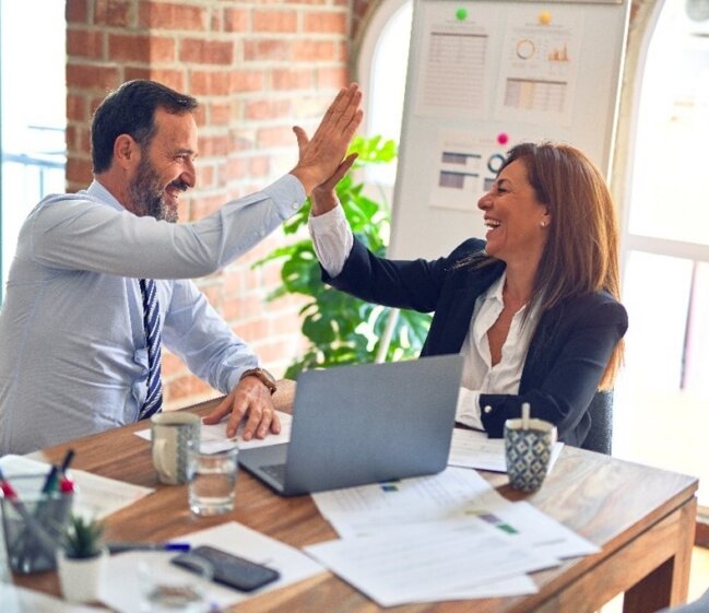 Two colleagues high-five over a desk with a laptop, papers, and mugs in a bright office featuring a brick wall and charts on a whiteboard.