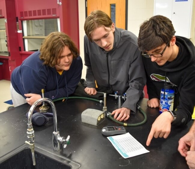 Three students in a science lab study an experimental setup with hoses, valves, and a digital meter on a black lab bench.