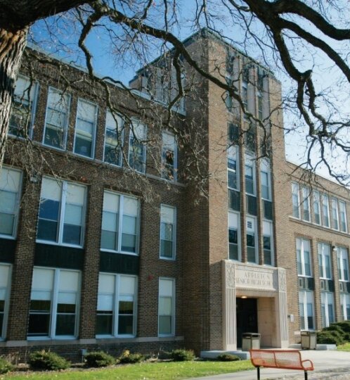 Brick campus building with a tall central entry and many windows, seen from behind a large tree trunk with bare branches; courtyard with benches.