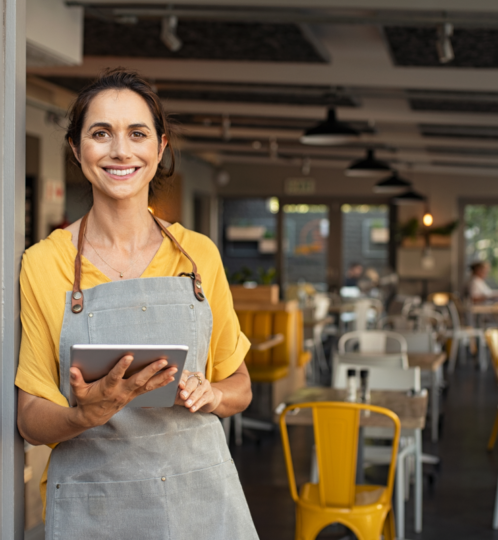 A person in a yellow shirt and gray apron stands by a doorway, holding a tablet and smiling in a busy cafe.