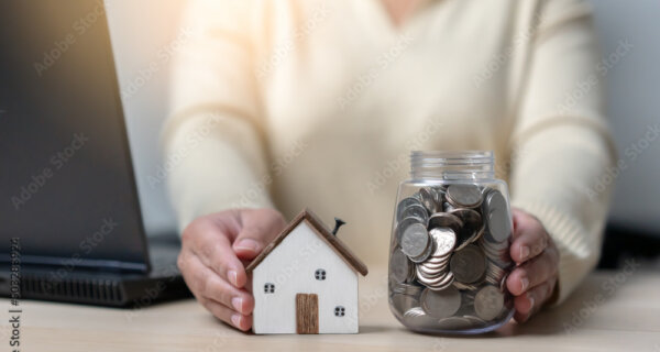 Person in a light sweater sits at a desk with a small wooden house model and a jar full of coins, beside a laptop, symbolizing saving or budgeting.
