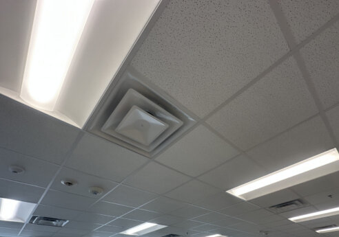 Office ceiling with suspended grid tiles, fluorescent light panels, and a square HVAC vent; windows along the wall show trees and parked cars outside.