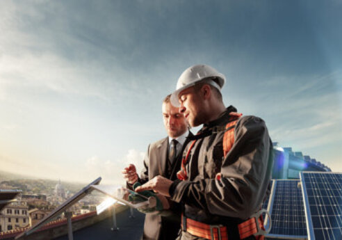 Two professionals on a rooftop with solar panels, one wearing a hard hat and safety gear, the other in a suit, inspecting a tablet.