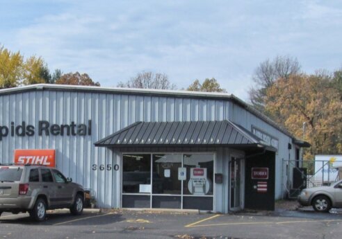Blue-gray metal building with a Rapids Rental sign and glass entry; two SUVs parked in front, autumn trees in the background.