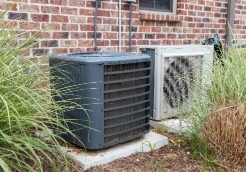 Two outdoor HVAC condenser units on concrete pads beside a brick wall, one dark gray with a large front grille and the other light gray with a round grill, with tall grasses nearby.
