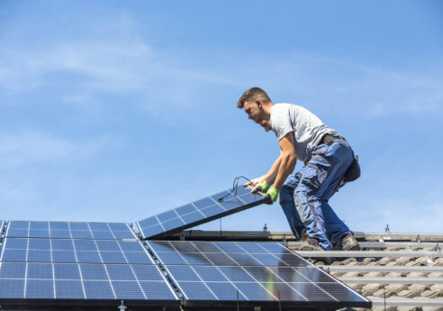 A worker on a roof connects a solar panel to a photovoltaic array, wearing gloves and a tool belt under a clear blue sky.
