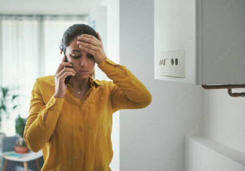 A person in a yellow shirt stands indoors, talking on a cell phone while resting a hand on their forehead in a stressed pose.