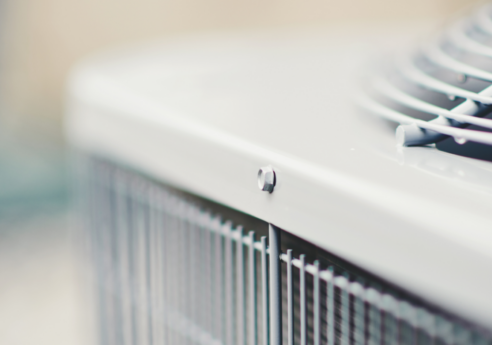 Close-up of an outdoor HVAC condenser: metal housing, top circular fan grille, and vertical cooling fins with a blurred background.