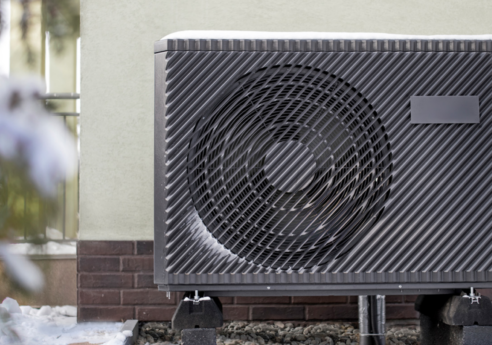Outdoor HVAC condenser unit with a diagonal striped grill and large circular fan, mounted on blocks beside a pale green wall, with snow on the ground.