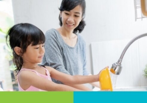 A child in pink uses a yellow spray nozzle at a bright kitchen sink while an adult smiles and watches nearby.
