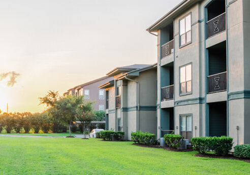 Sunset over a beige apartment complex with balconies on the right, a green lawn in the foreground, and trees along a fence on the left.