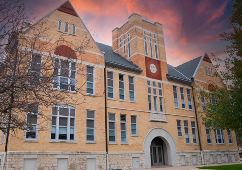 Brick campus building with an arched entry, tall clock tower, and many windows; stone base, leafless trees, and a pink-orange sunset sky.