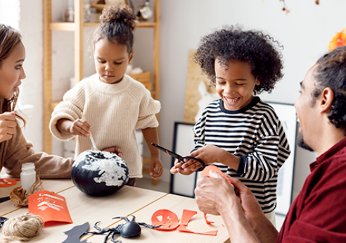 A diverse group of four around a sunlit table making crafts; a globe, scissors, glue, yarn, and red paper on the table as they smile and work together.