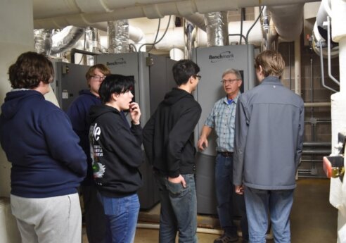 An older person in a plaid shirt explains to a group of young people in a mechanical room with large gray Benchmark machines and exposed ductwork.