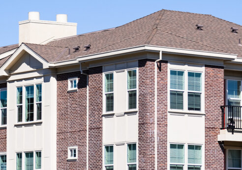 Brick apartment building with beige trim, windows with blinds, a small balcony with a black railing, and a brown shingle roof under a blue sky.