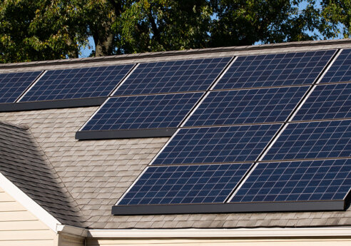 Residential roof with multiple dark blue solar panels mounted on shingles under a clear sky.