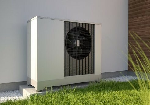 Outdoor air conditioner condenser unit on a small concrete pad beside a white wall, with a visible fan behind a grille, surrounded by gravel and green grass.